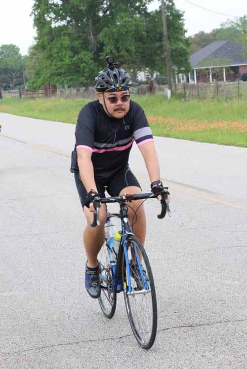 - Male cyclist riding on a paved road with trees and houses in the background.