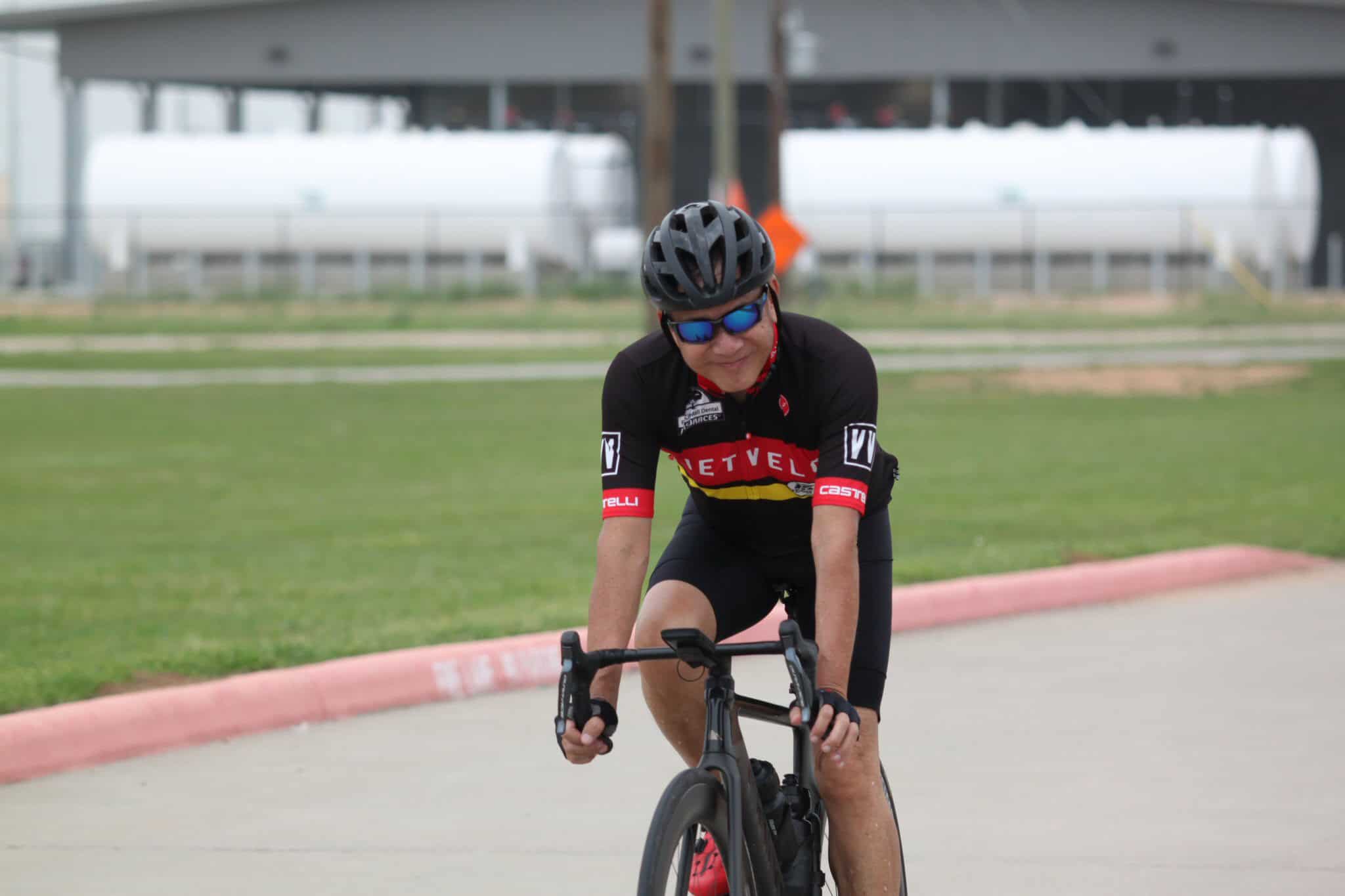 Bright cyclist in racing gear on black bike, wearing helmet and sunglasses, riding on smooth concrete track.