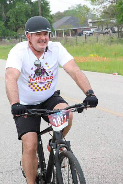 Smiling man riding a mountain bike with race number 1837, participating in outdoor cycling event.