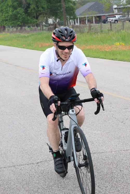 High-performance cyclist wearing helmet and sunglasses riding on a paved road during daytime.