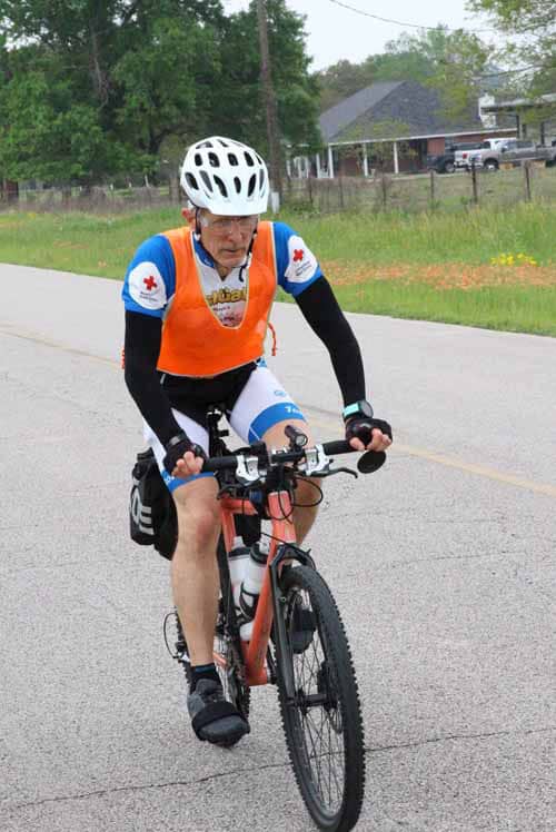 Older man cycling on a rural road during daytime wearing safety gear and athletic clothing.