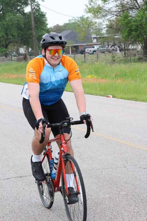 Bright cyclist on a road bike wearing a blue and orange jersey, black helmet, and sunglasses during daytime ride.