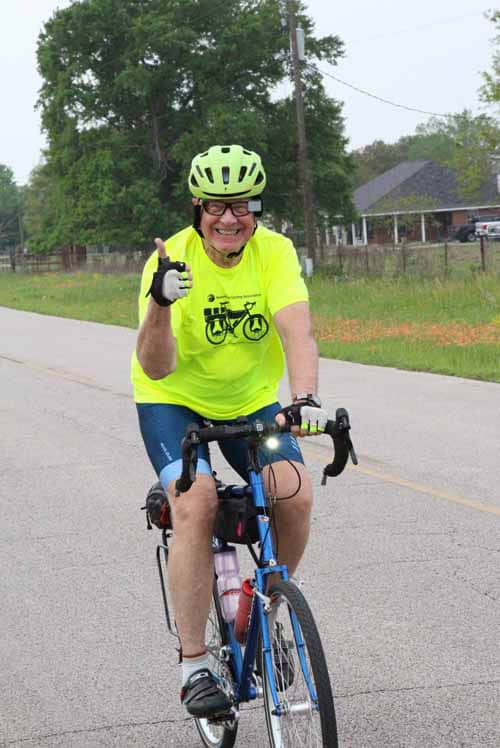 Bright yellow cyclist jersey, smiling man riding bike on scenic road, green trees in background, promoting outdoor biking.