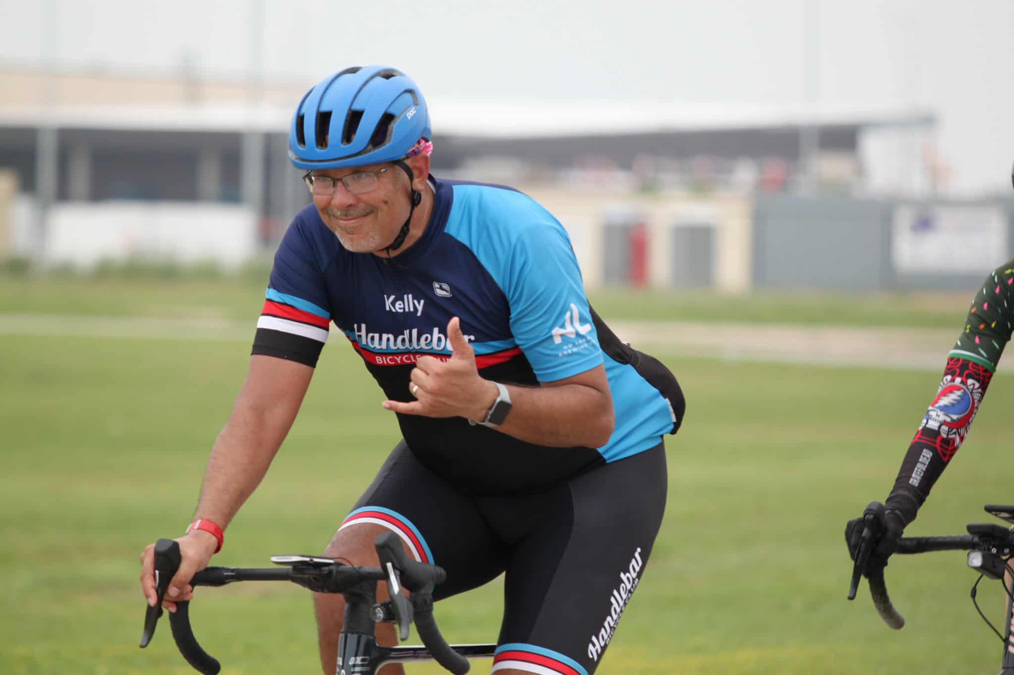Smiling male cyclist wearing a blue helmet and Handlebar Bicycle jersey, giving a thumbs-up during a ride.