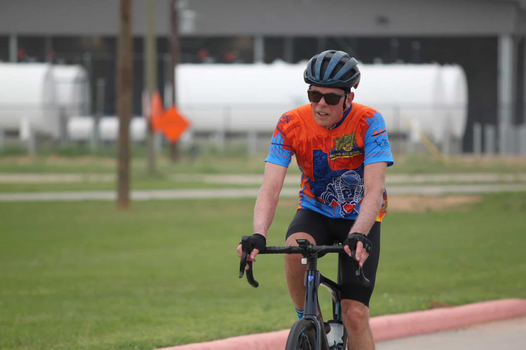 Bright orange and blue cycling jersey, male cyclist wearing a helmet and sunglasses riding a bike.