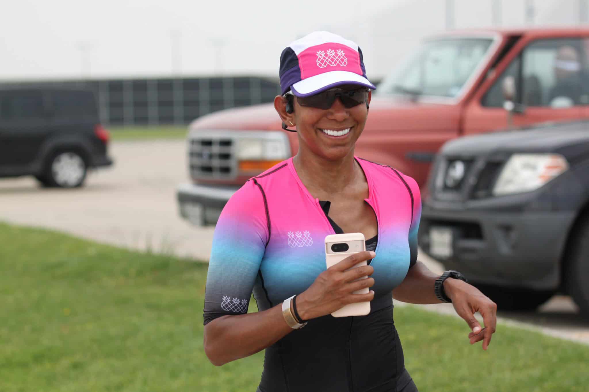 Bright smiling female cyclist in vibrant Pink and Blue athletic jersey, holding smartphone during outdoor ride.