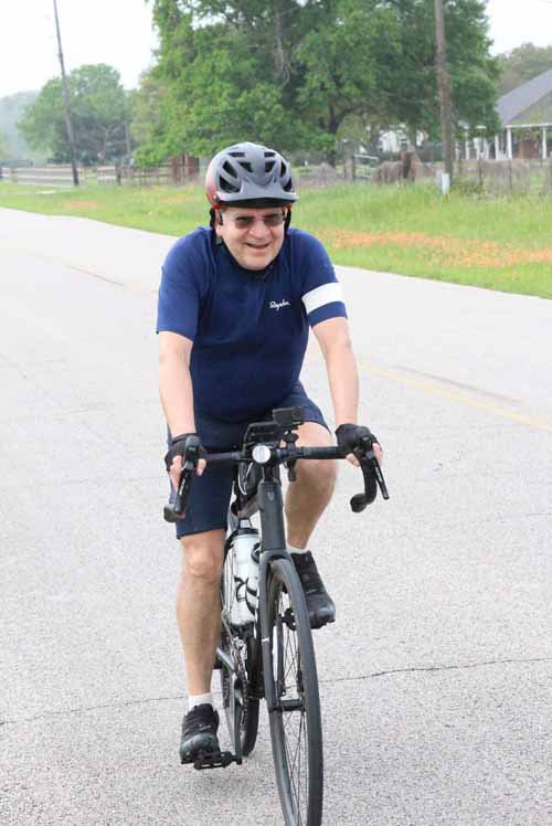 Smiling man cycling on open road during daytime, wearing helmet and sports gear, enjoying outdoor fitness.