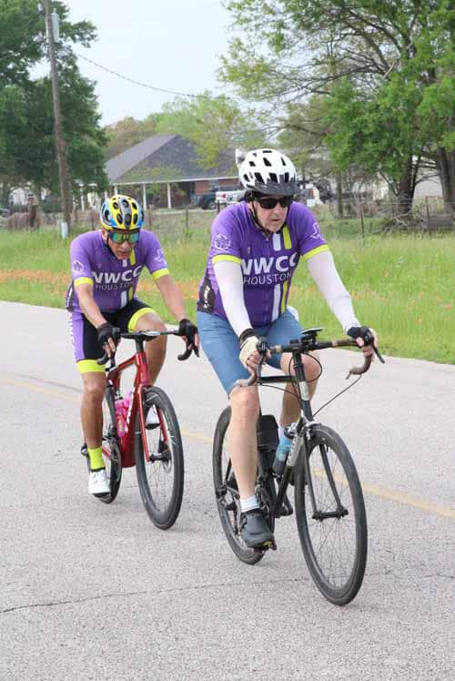 Bright purple cycling jerseys, two cyclists riding together on a rural road during daytime.
