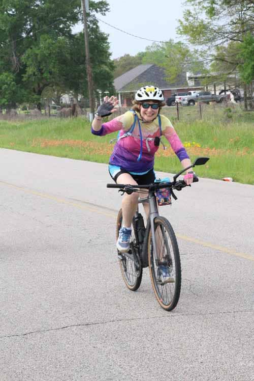 Smiling woman riding a bicycle, waving, wearing helmet and colorful cycling gear outdoors.