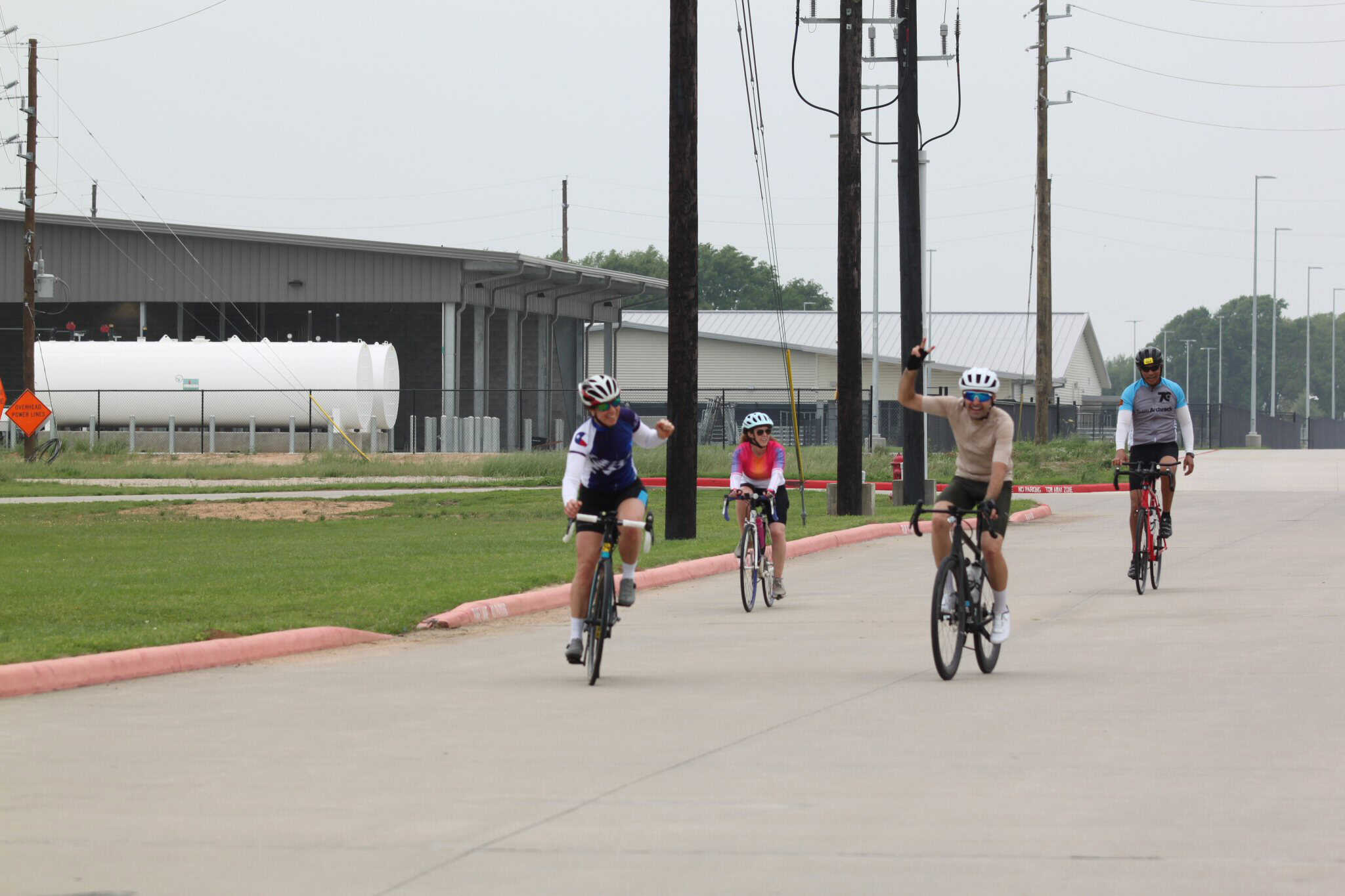 Cyclists enjoying a group ride on a paved path, wearing helmets and activewear, with power lines and industrial buildings in the background.