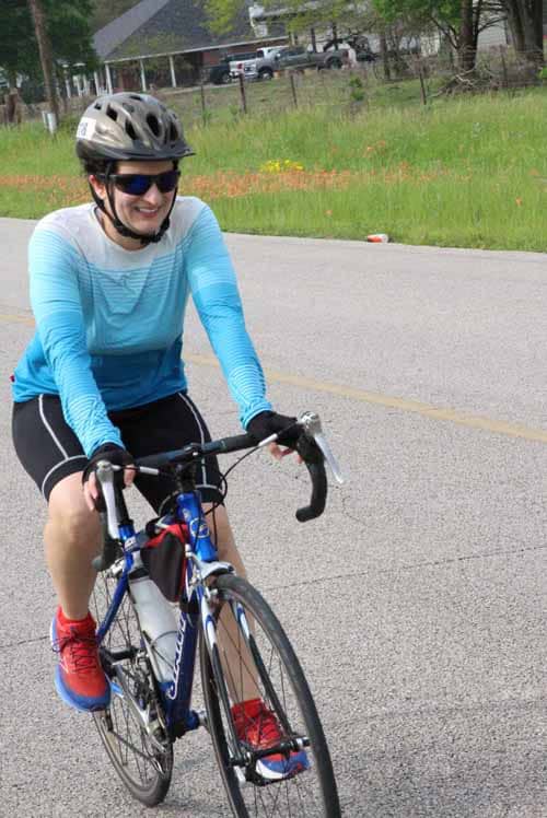 A woman riding a blue bike on the road wearing a helmet and cycling gear, enjoying outdoor cycling.