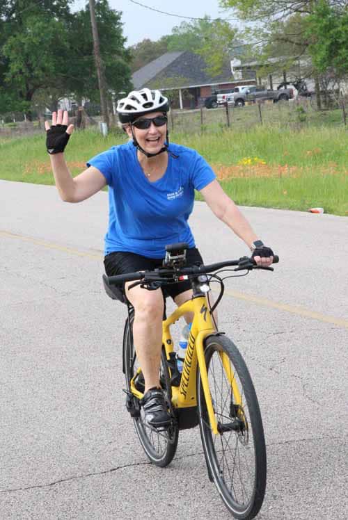 1. Woman cycling on bike wearing helmet and sunglasses, smiling and waving, outdoors in a scenic area.