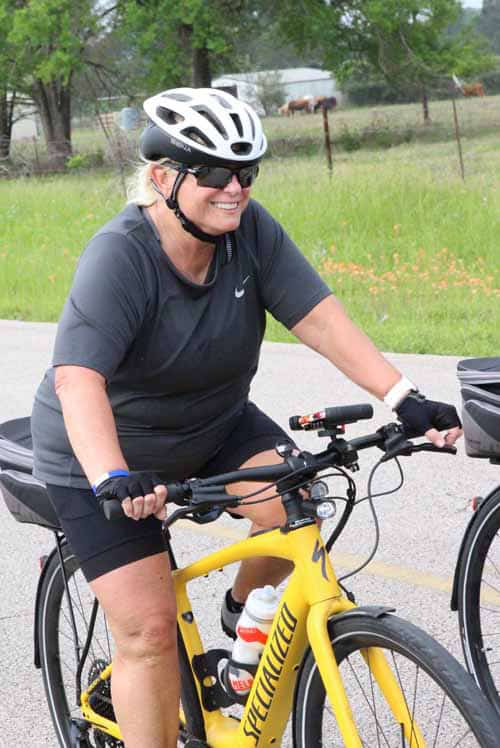 Smiling woman cycling on a sunny road with green trees and horses in the background.