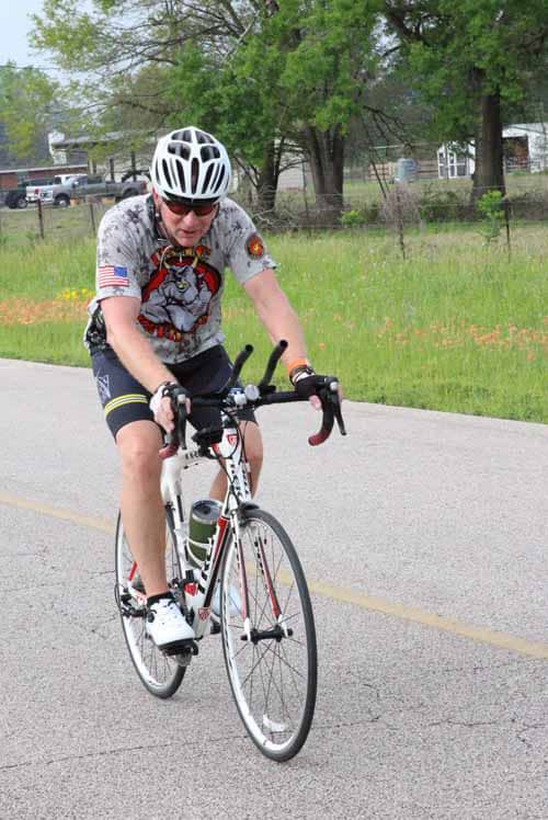 Bright cyclist riding on the road with greenery and houses in background, promoting Bluebonnet Express biking event.