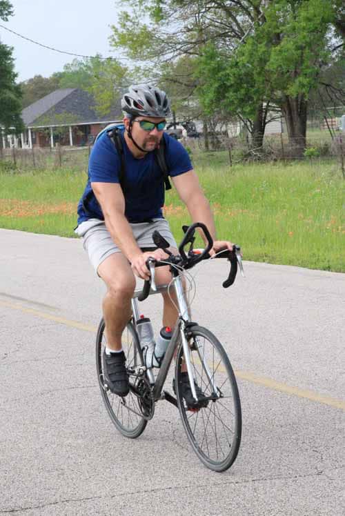 1. Man riding a bicycle along a rural road with trees and houses in the background.
