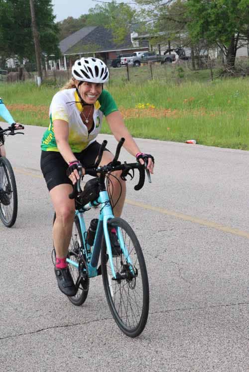 Bright smiling woman riding a blue mountain bike wearing a white helmet on a paved road.