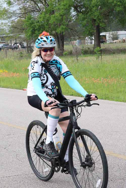 Woman cyclist riding a black bike on a rural road in colorful cycling gear and helmet.