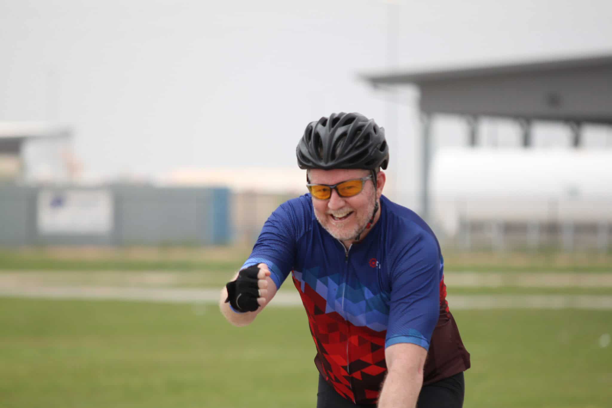 Happy man riding a bicycle with Bluebonnet Express cycling jersey, outdoor bike path, Texas scenery, cycling event, smiling cyclist, athletic activity, outdoor sports.