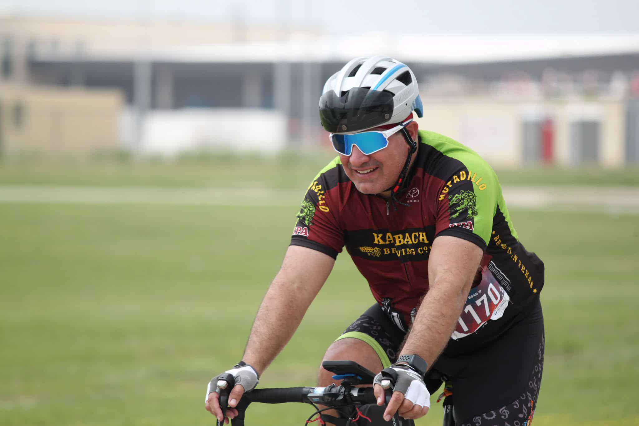 Brightly clothed cyclist wearing helmet and sunglasses riding on green outdoor trail.