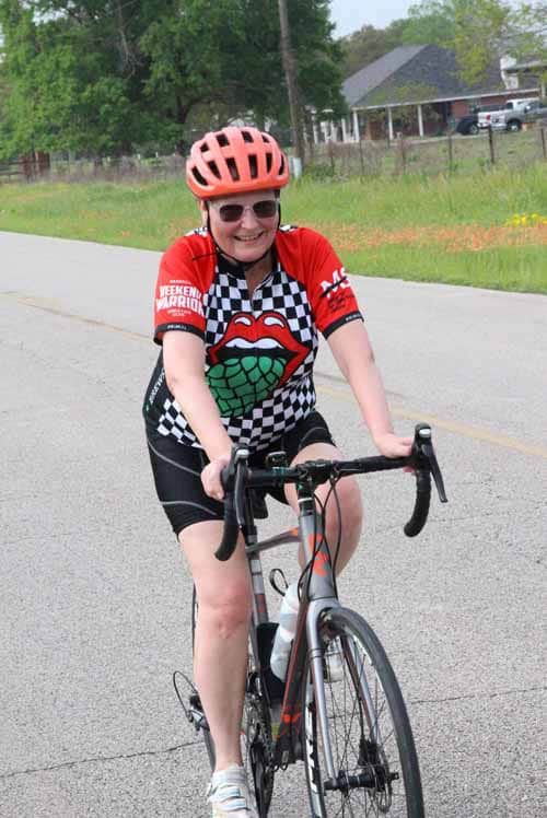 Pink bike helmet, smiling cyclist in checkered Jesus Lizard jersey, riding on pavement.