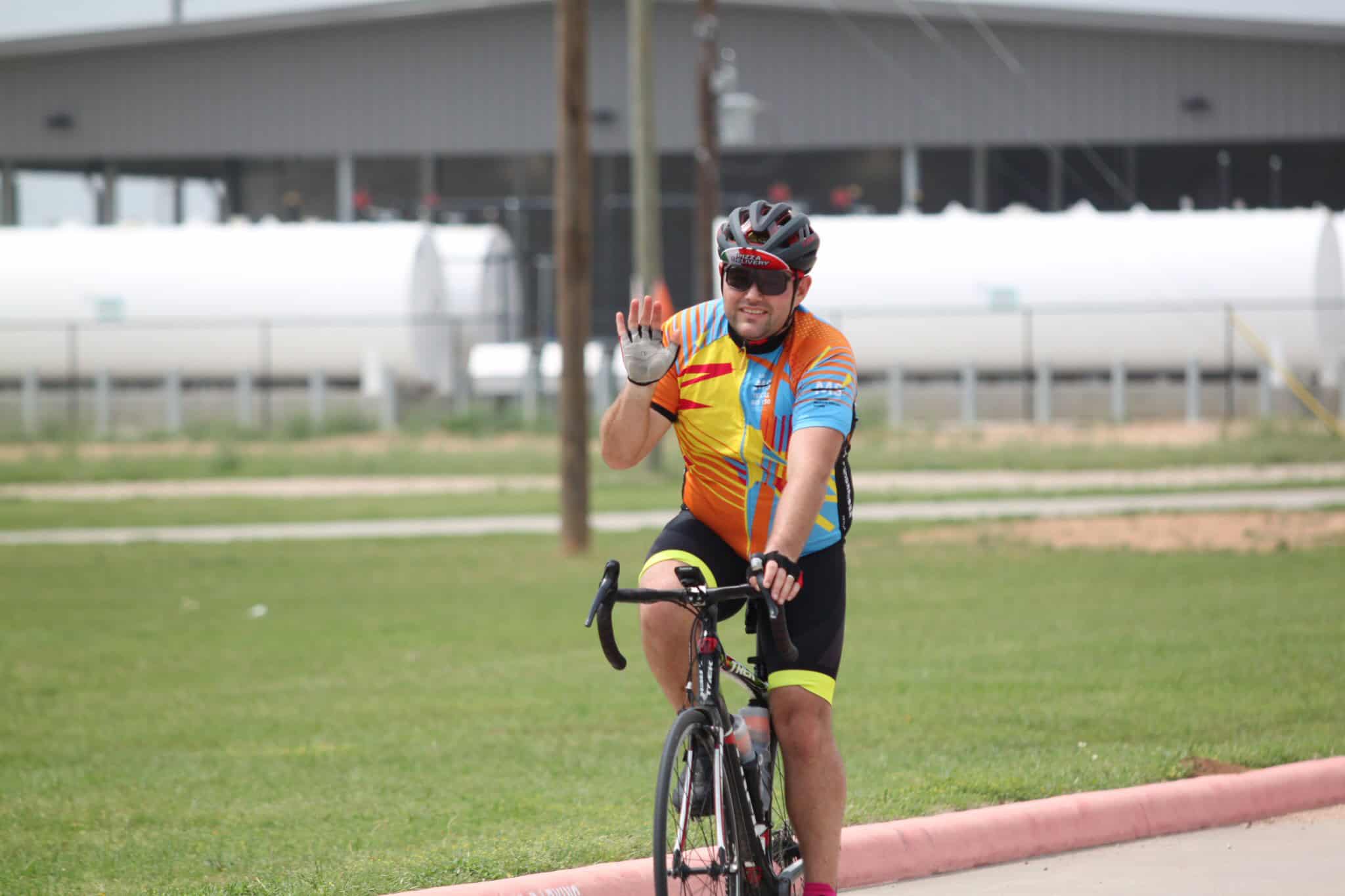 Vibrant cyclist waving, wearing colorful cycling gear, on a sunny day in a park near industrial buildings.