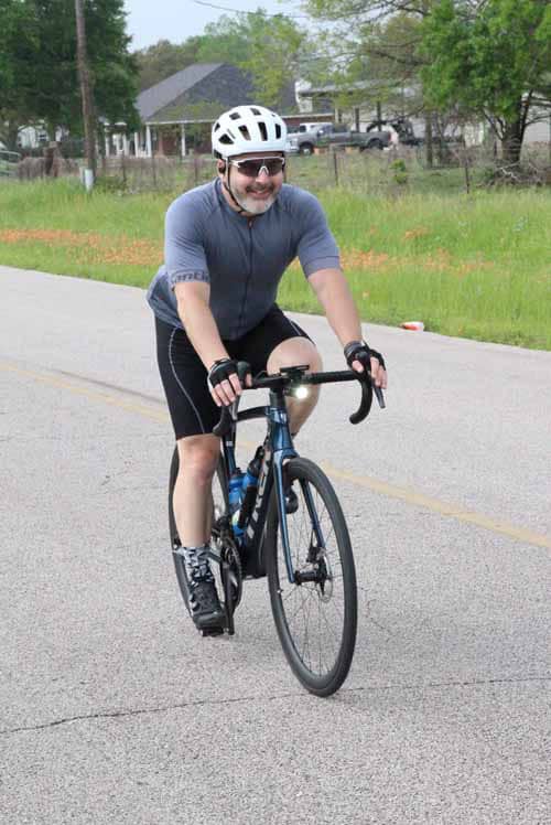 Smiling man riding a bike on the road with greenery in background, wearing helmet and athletic gear.