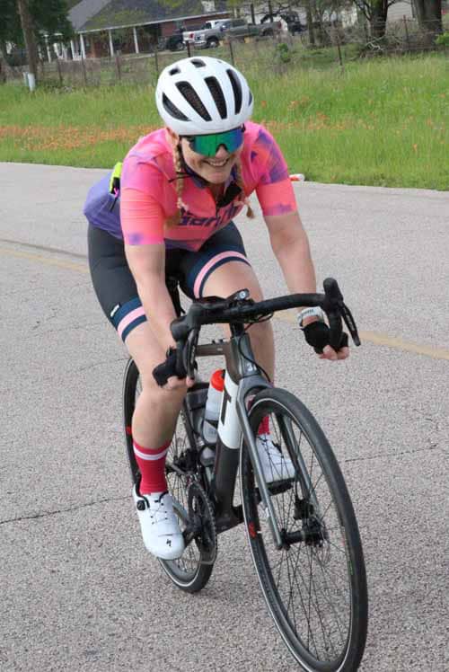 Vibrant female cyclist wearing pink and purple gear riding a road bike on pavement for Bluebonnet Express.