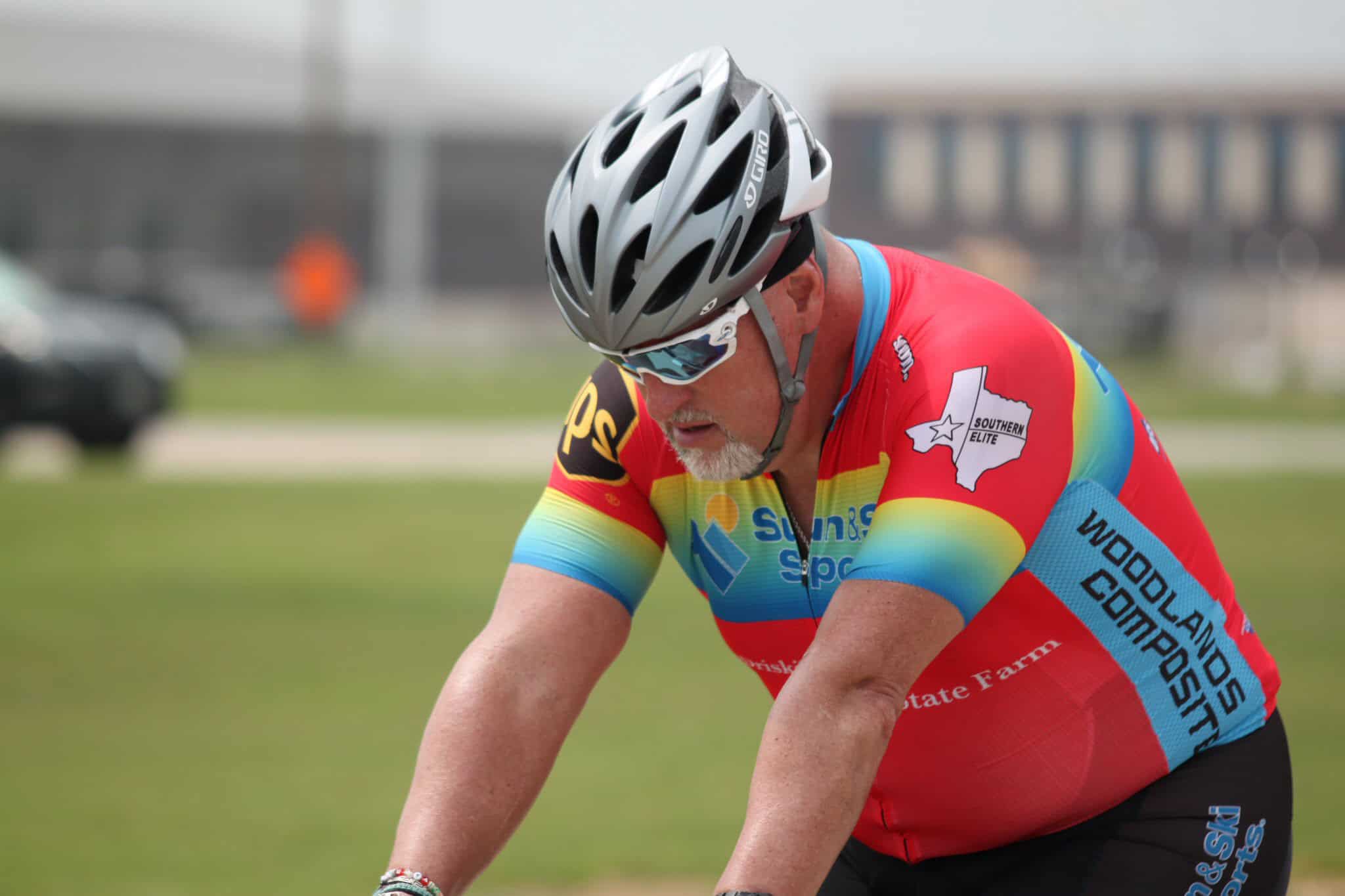 Velo cyclist wearing colorful jersey and helmet, riding high-speed on a road.