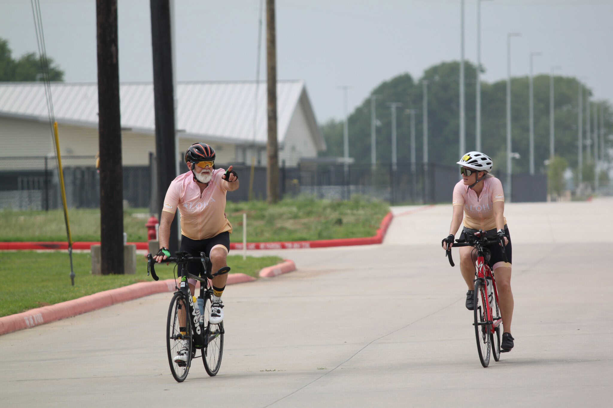 Older man biker pointing at young cyclist while riding on city street, both wearing helmets, promoting bike safety and family fun.