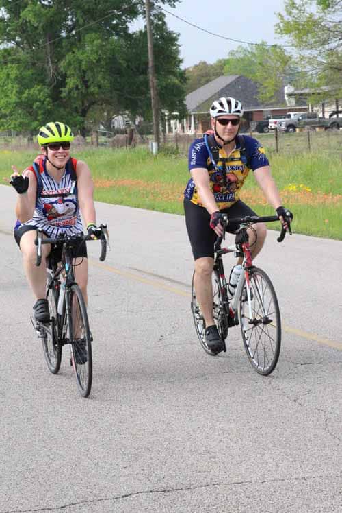 Brightly dressed cyclists enjoying a ride on a rural road with lush greenery and neighborhood houses in the background.