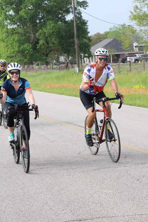 - Cyclists riding on scenic suburban road wearing helmets and colorful cycling jerseys for Bluebonnet Express event.