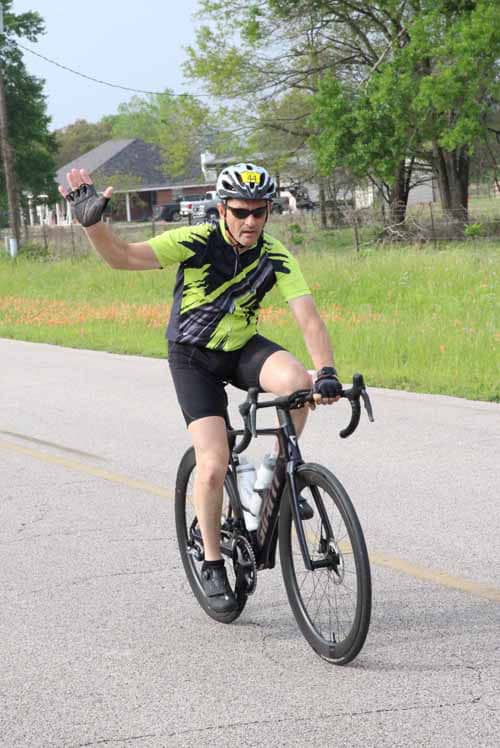 Rider waving on a road bike outdoors with green trees and houses in background.