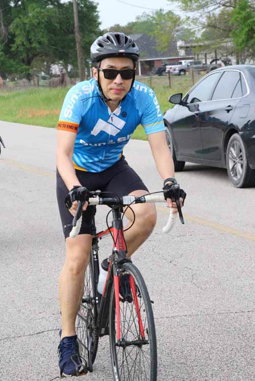 Bike rider wearing Bluebonnet Express cycling jersey on road with black car in background.