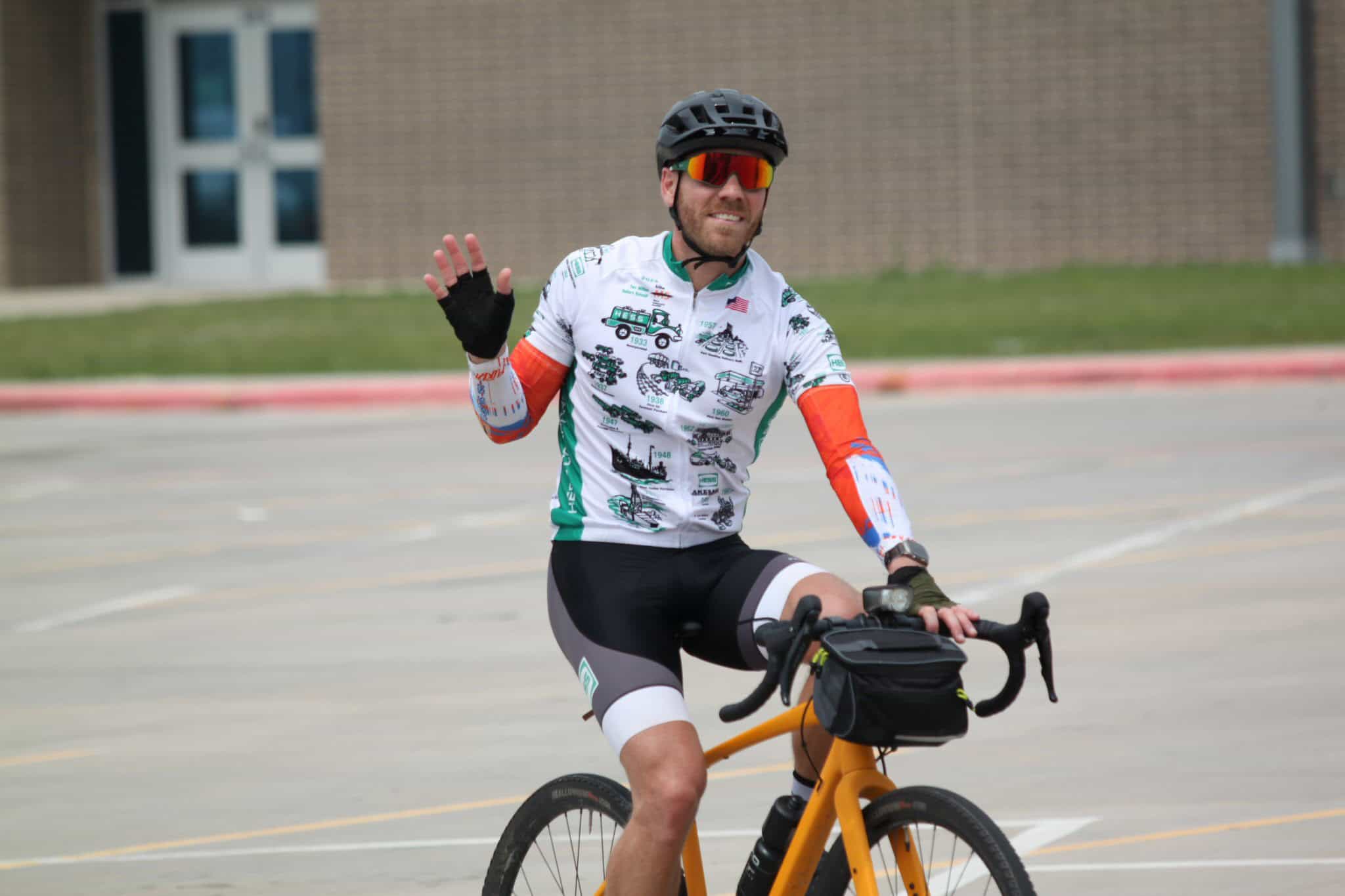 Rider waving on a yellow bicycle, wearing cycling gear, helmet, and sunglasses, during Bluebonnet Express event.