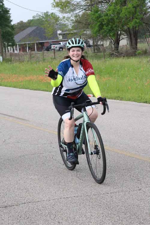 Bright smiling woman riding a bicycle on a rural road during daytime, wearing a helmet and sports gear.