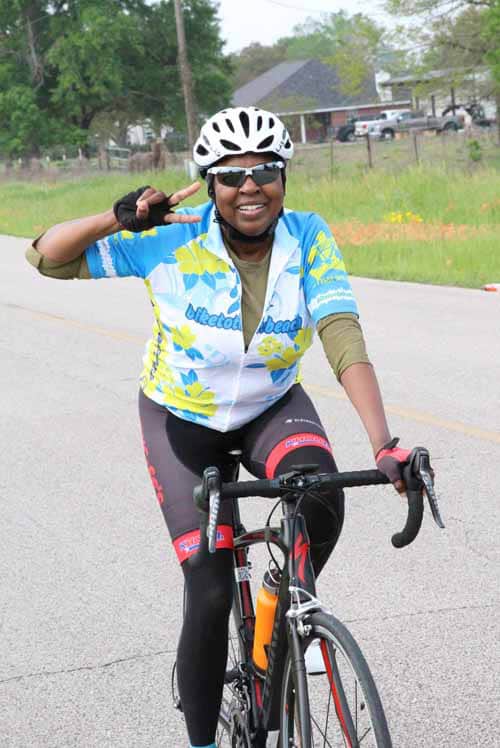 1. Woman cyclist wearing Bluebonnet Express jersey smiling and making a peace sign on a bike.