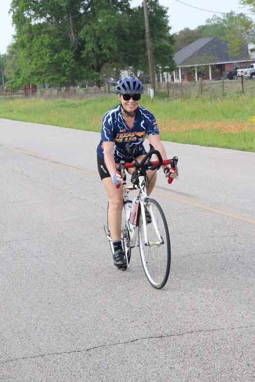 Bright female cyclist riding on rural road, wearing Bluebonnet Express cycling apparel, enjoying outdoor adventure.