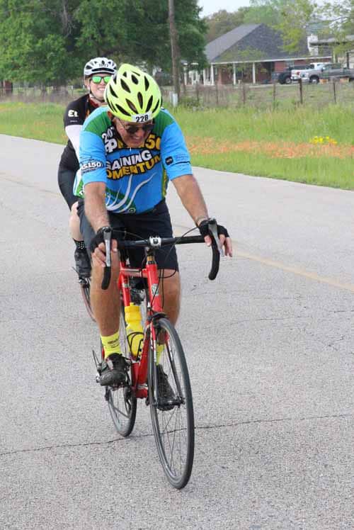 Bright blue and green cycling jersey, two male cyclists riding on a paved road during Bluebonnet Express event.