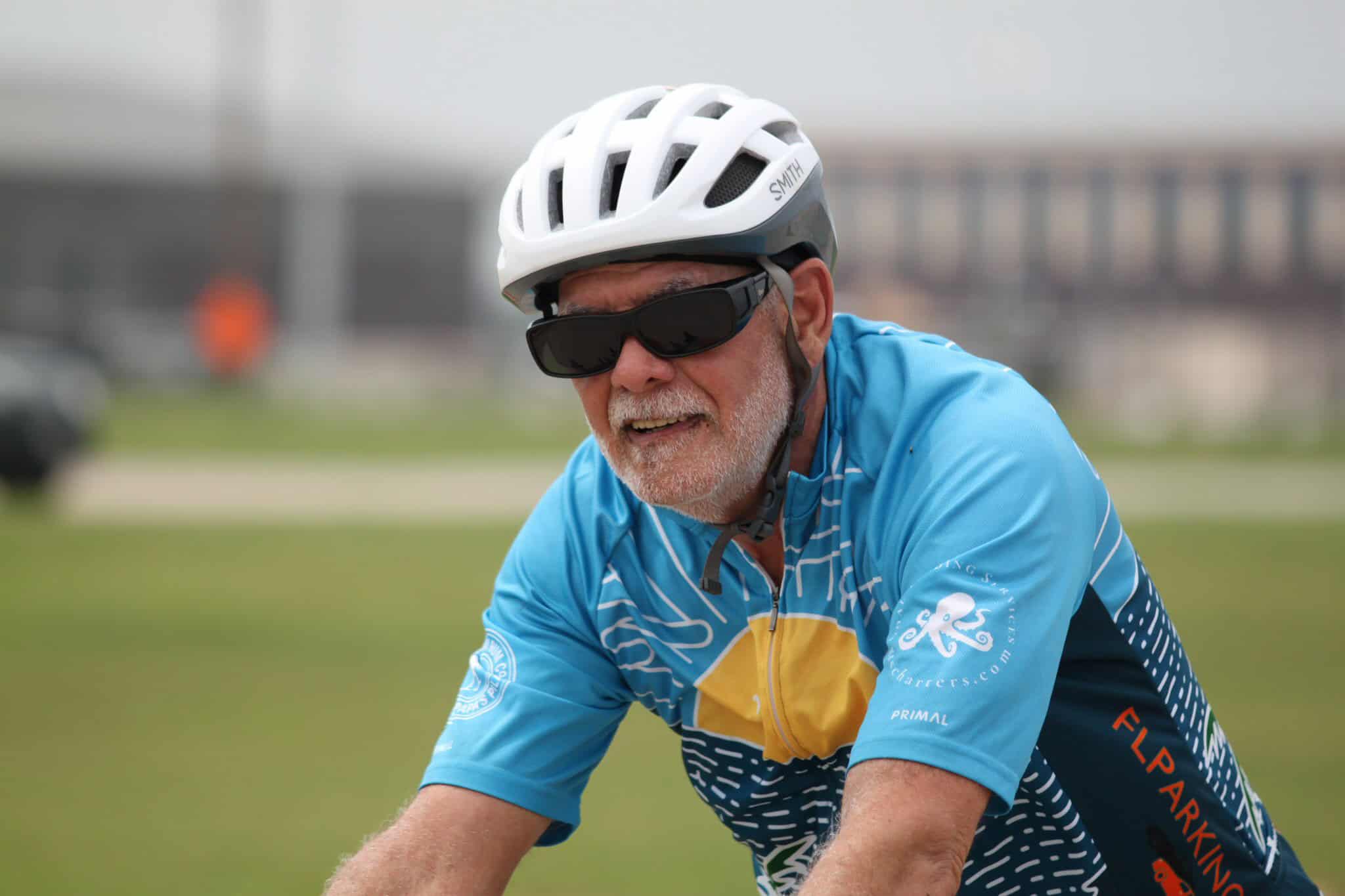 Older man biking in Bluebonnet Express cycling jersey, wearing helmet and sunglasses for safety and comfort.
