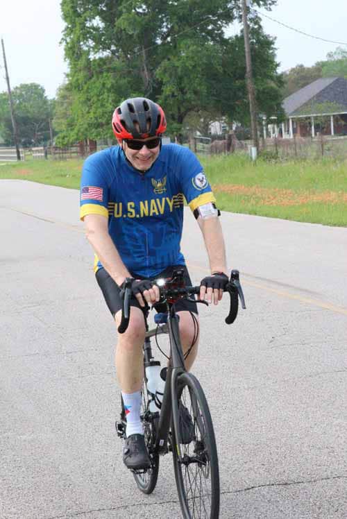 U.S. Navy cyclist riding on a rural road during daytime, wearing a blue uniform and helmet.