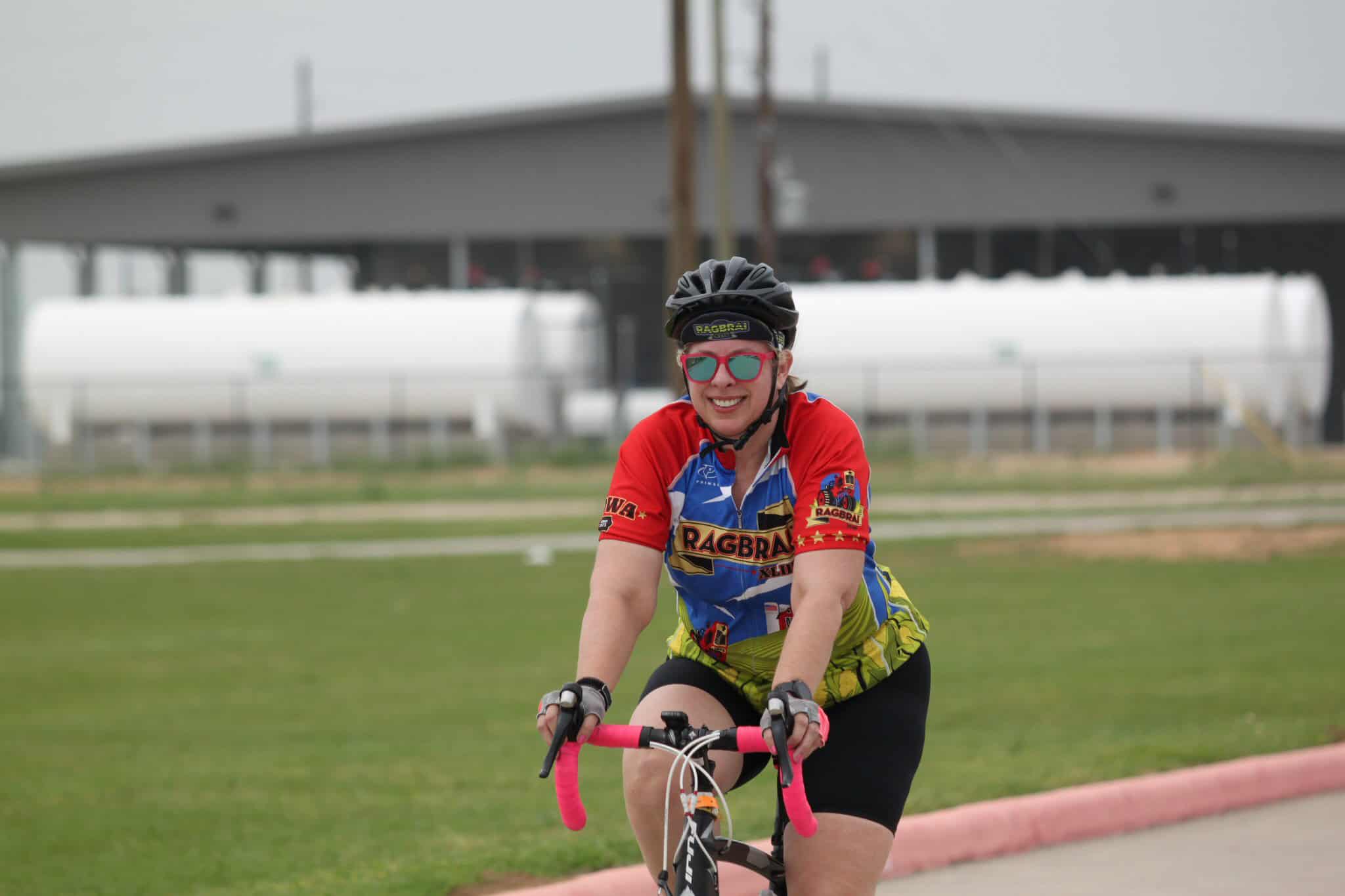 Vibrant female cyclist wearing RAGBRAI jersey and helmet riding bike outdoors on a cloudy day.