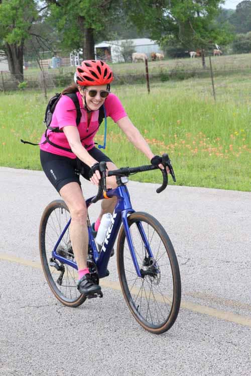 Bright female cyclist riding Bluebonnet Express bike on rural road with trees and fields in background.