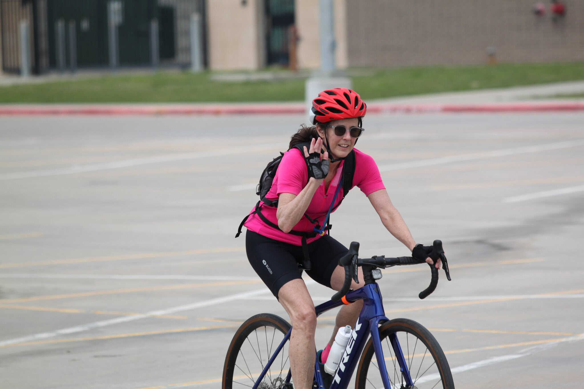 Bright pink cycling jersey with woman wearing helmet and sunglasses riding a blue bike in an open parking lot.