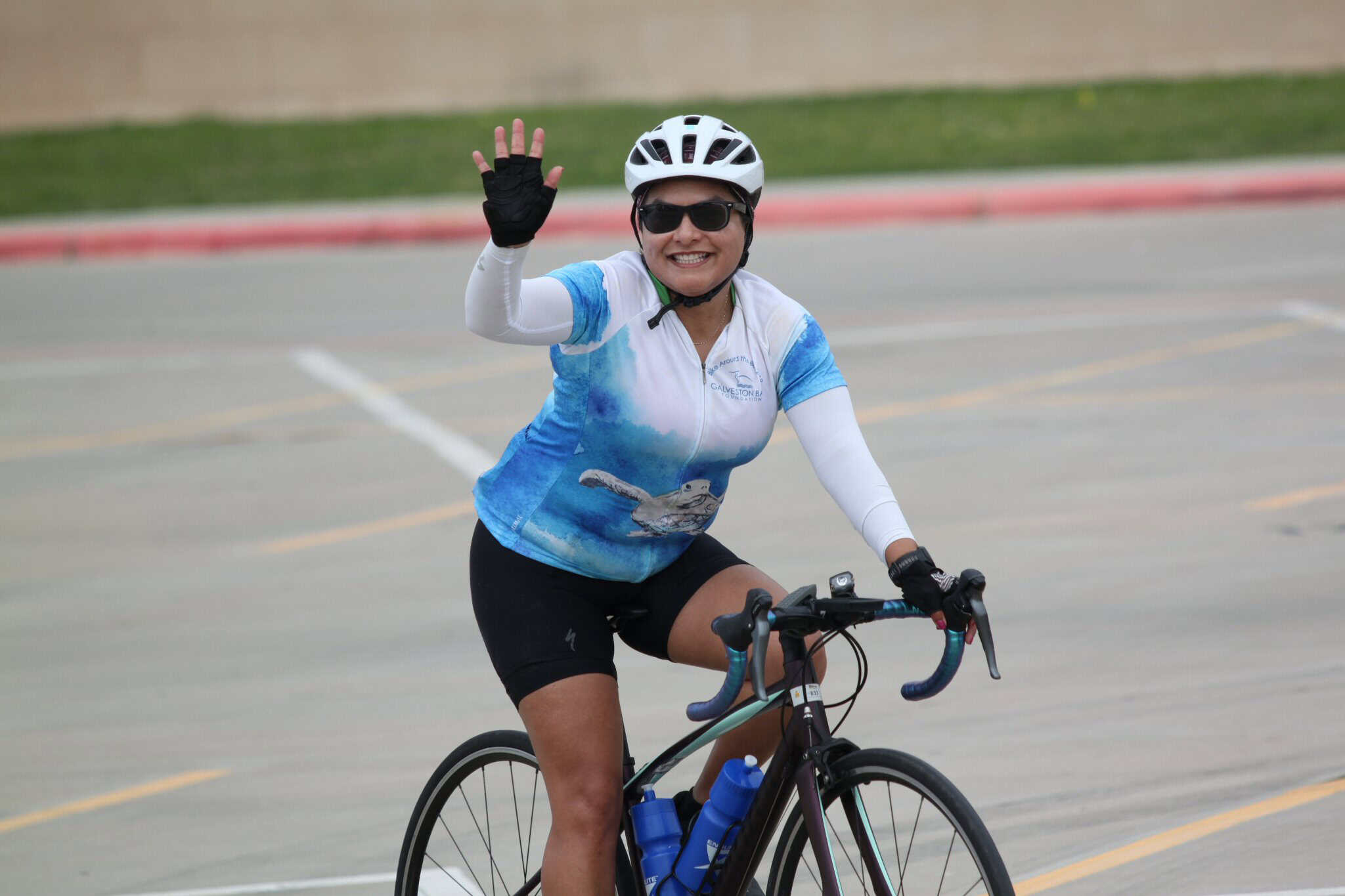 Happy cyclist riding Bluebonnet Express bike, waving at camera, outdoor scenic setting.