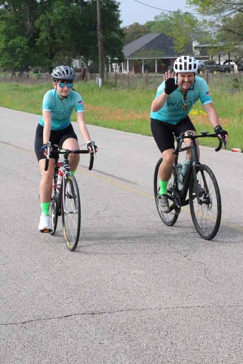 Riders wearing Bluebonnet Express jerseys cycling on a scenic road with trees and houses.
