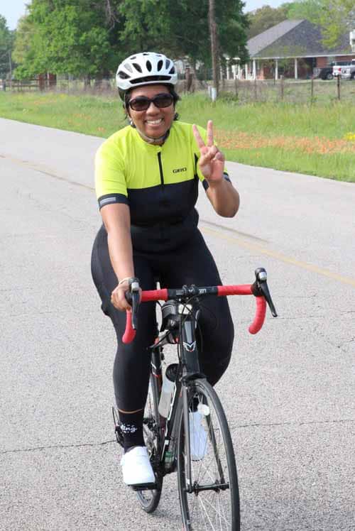 Bright cyclist in yellow and black jersey riding a road bike on quiet street, making a peace sign.