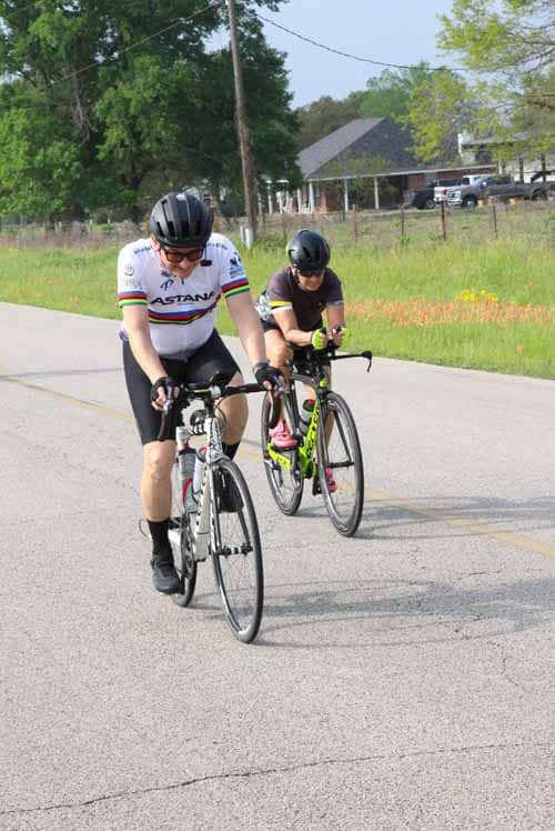Riders cycling on a sunny rural road in Bluebonnet Express event, outdoors, green scenery, promoting bike riding and outdoor fitness.