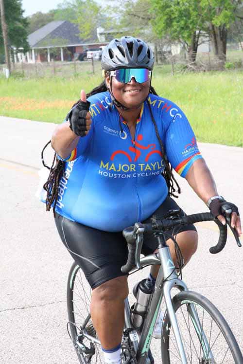 Vibrant woman cyclist wearing blue jersey, helmet, and sunglasses, giving a thumbs-up during riding outdoors.