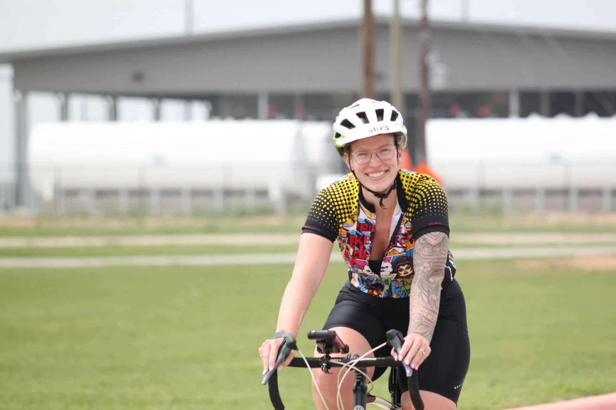 Vibrant woman cyclist smiling, wearing a colorful jersey and helmet, riding on a green field for Bluebonnet Express.
