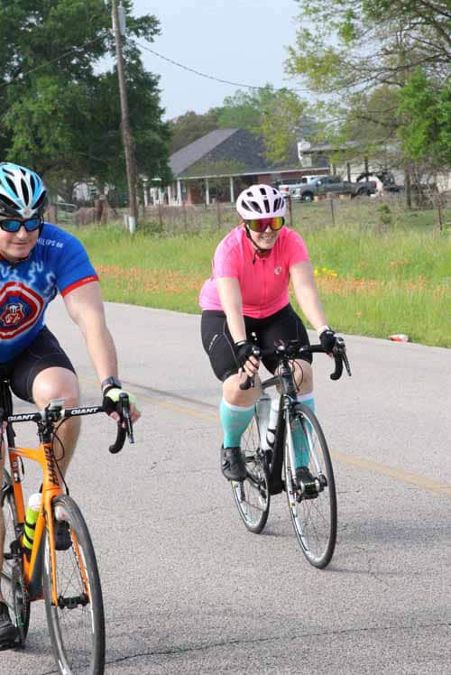 - Bright female cyclist wearing pink and white helmet riding on open road with green trees around.